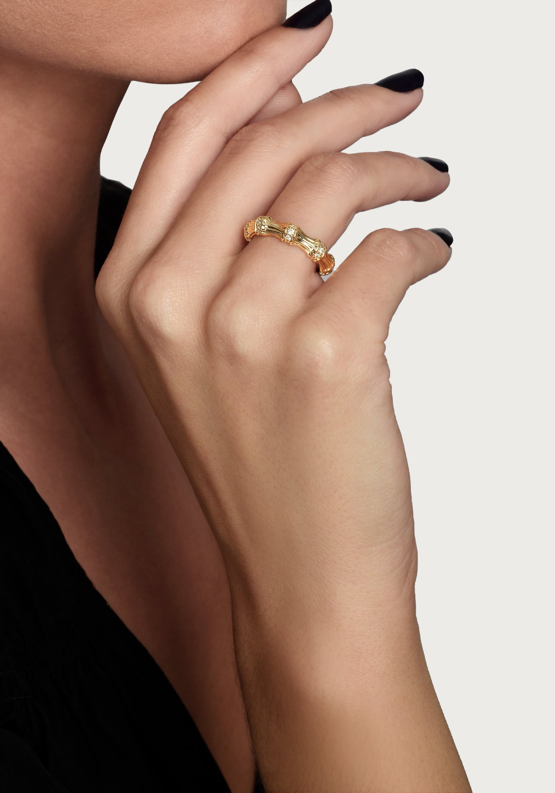 A close-up of a hand with black nail polish wearing the Anabel Aram Bamboo Single Ring, an elegant 18k gold design, held near the chin against a plain background.