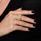 A hand with dark polished nails rests on black fabric, showcasing the Anabel Aram Bamboo Stack Ring—a thick, 18k gold dipped band with an intricate bamboo-inspired design and pavé crystal accents on the middle finger.