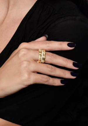 A hand with dark polished nails rests on black fabric, showcasing the Anabel Aram Bamboo Stack Ring—a thick, 18k gold dipped band with an intricate bamboo-inspired design and pavé crystal accents on the middle finger.
