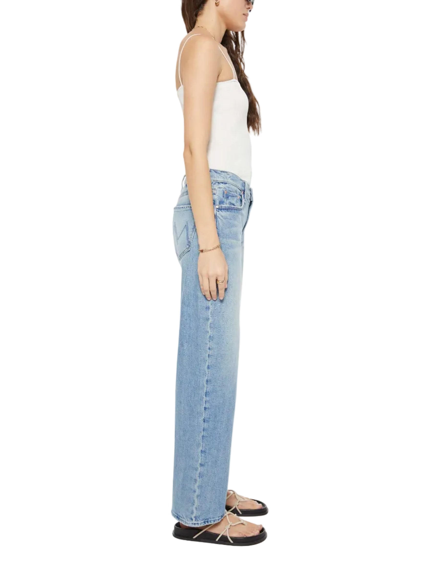 Woman in profile wears a white tank top, Mother Mid Rise Zip Rambler Skimp light blue denim wide straight leg jeans, and black flat sandals with white straps.