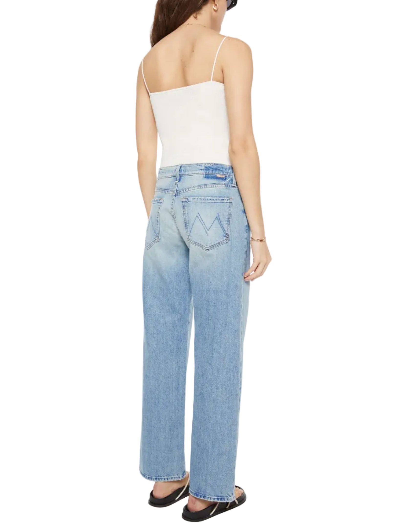 A woman in a white tank top, black sandals, and Mother’s Mid Rise Zip Rambler Skimp light blue denim jeans stands facing away from the camera.