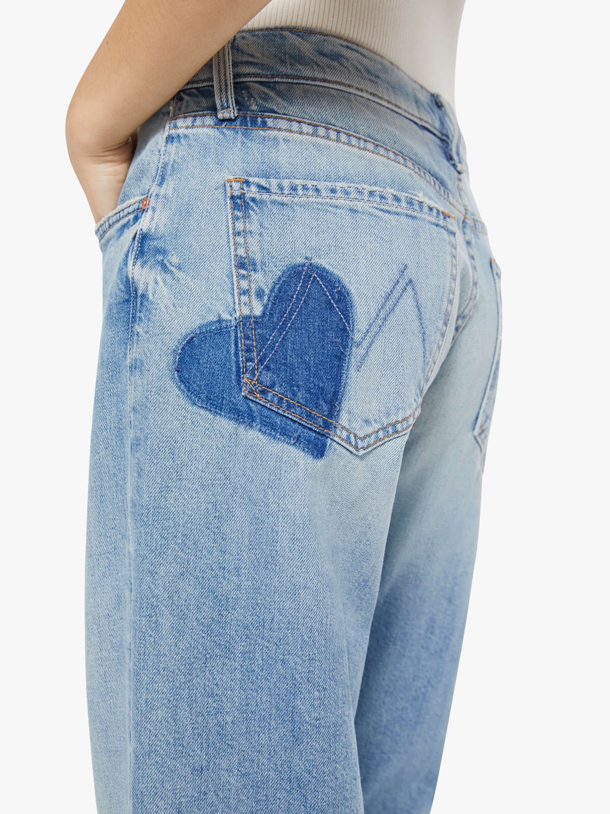 A close-up of a person wearing Mother’s Mid Rise Zip Rambler Skimp jeans—wide, straight leg, light blue denim with a dark blue heart on the back pocket.