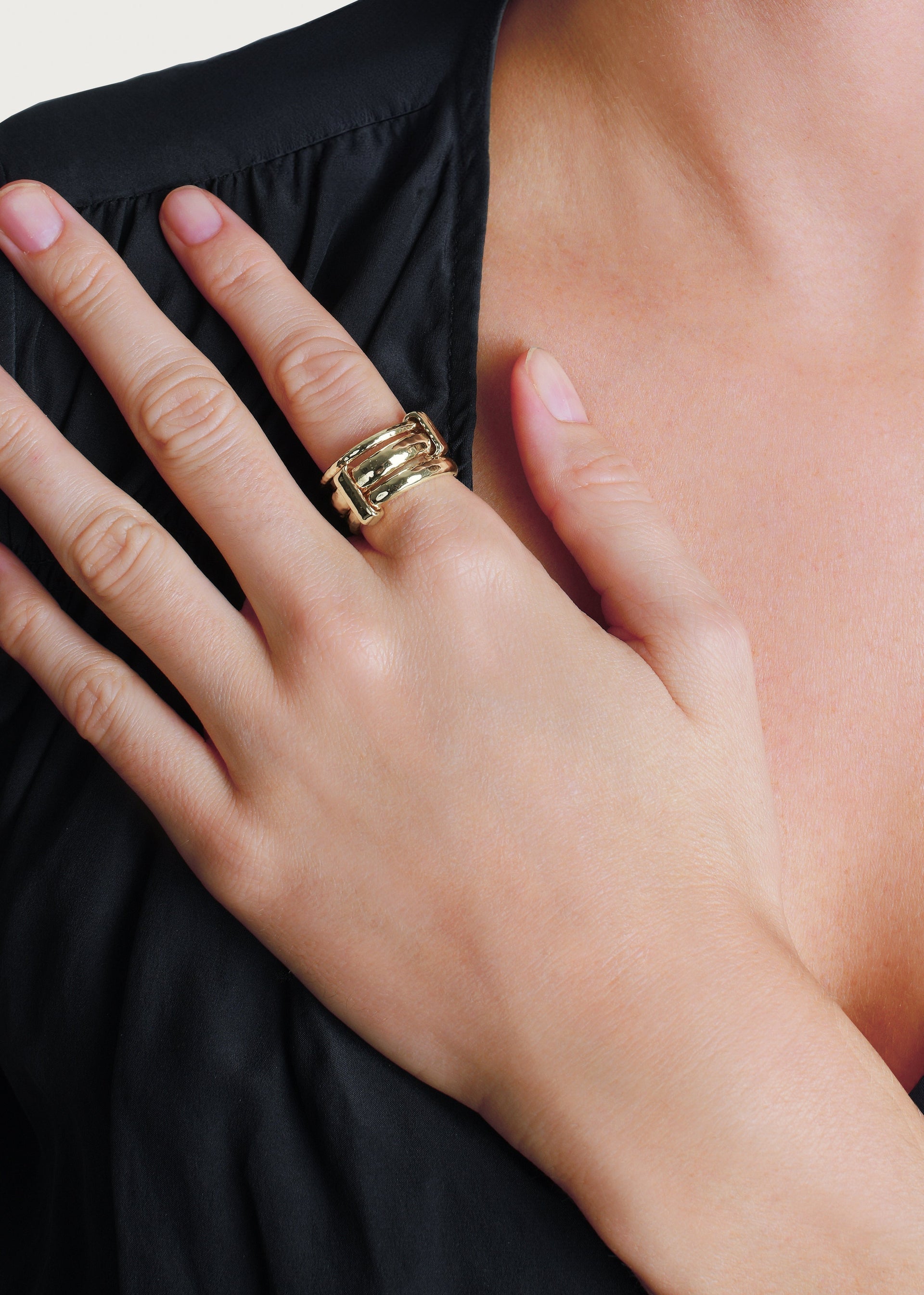 A person wears the Anabel Aram Farrier Triple Connected Ring—a large, gold, hammered ring—on their middle finger, resting their hand on their chest while dressed in black.