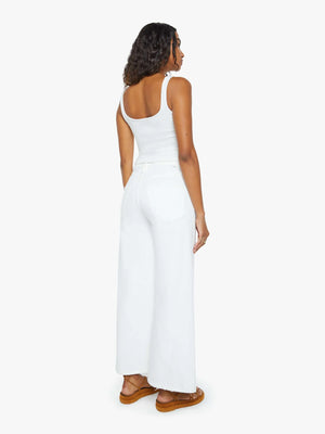 A woman stands facing away, wearing Mother’s The Ditcher Roller Zip Ankle jeans with a white tank top and brown sandals against a plain white background.