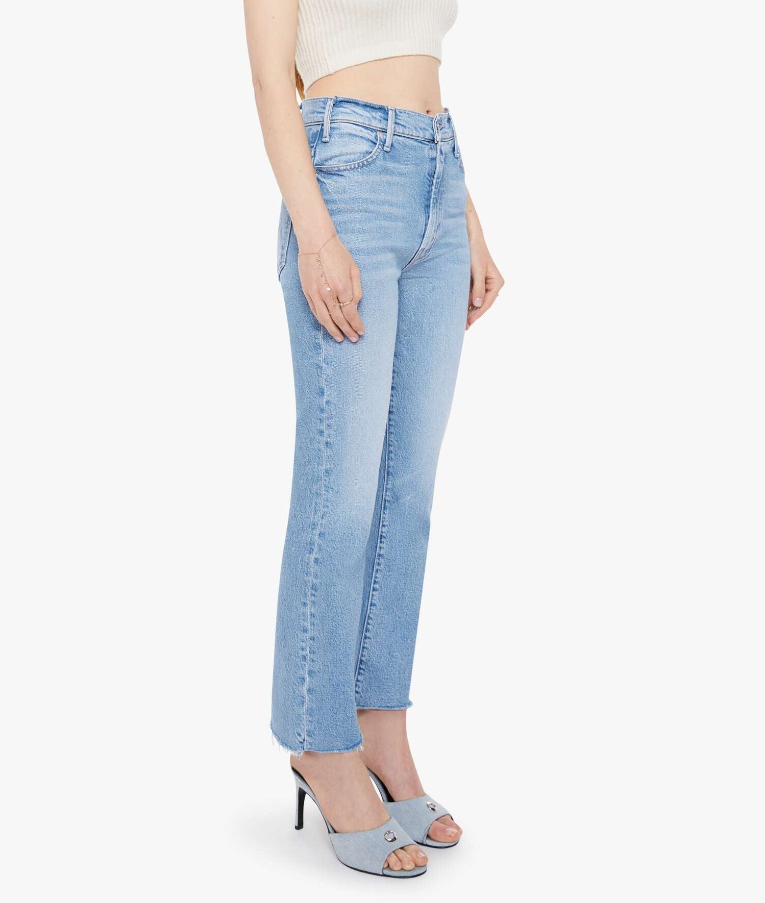 A person wears Mother’s The Hustler Ankle Fray high-waist flare jeans with a raw hem, styled with a white sleeveless crop top and light blue heeled sandals, standing against a plain white background.