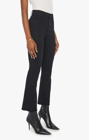 A person wearing Mother’s The Hustler Ankle Fray high-waist flare jeans in stretch denim, paired with a white top and black heeled ankle boots, stands against a plain white background.