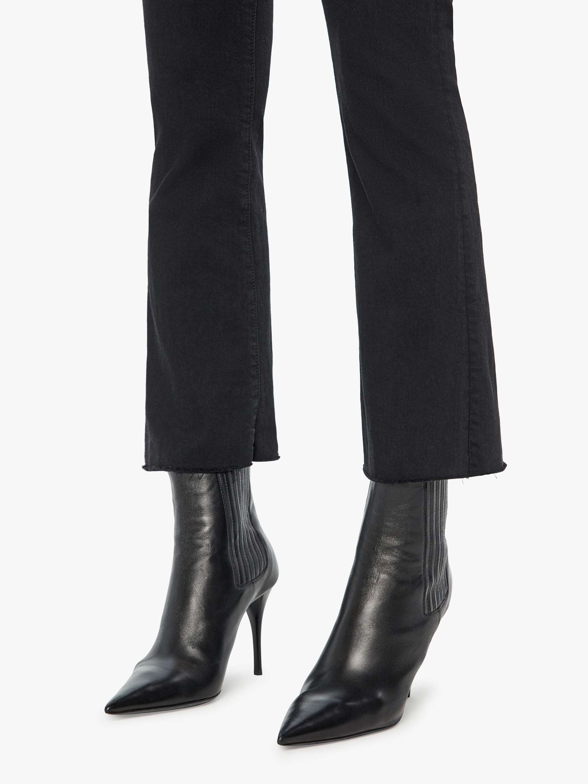 Person wearing black pointed-toe heeled ankle boots and Mother’s The Hustler Ankle Fray high-waist flare jeans with a raw hem against a white background.
