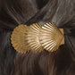 The Jennifer Behr Siren Barrette, shaped like two gold seashells, holds back dark brown hair securely with a French barrette clasp.
