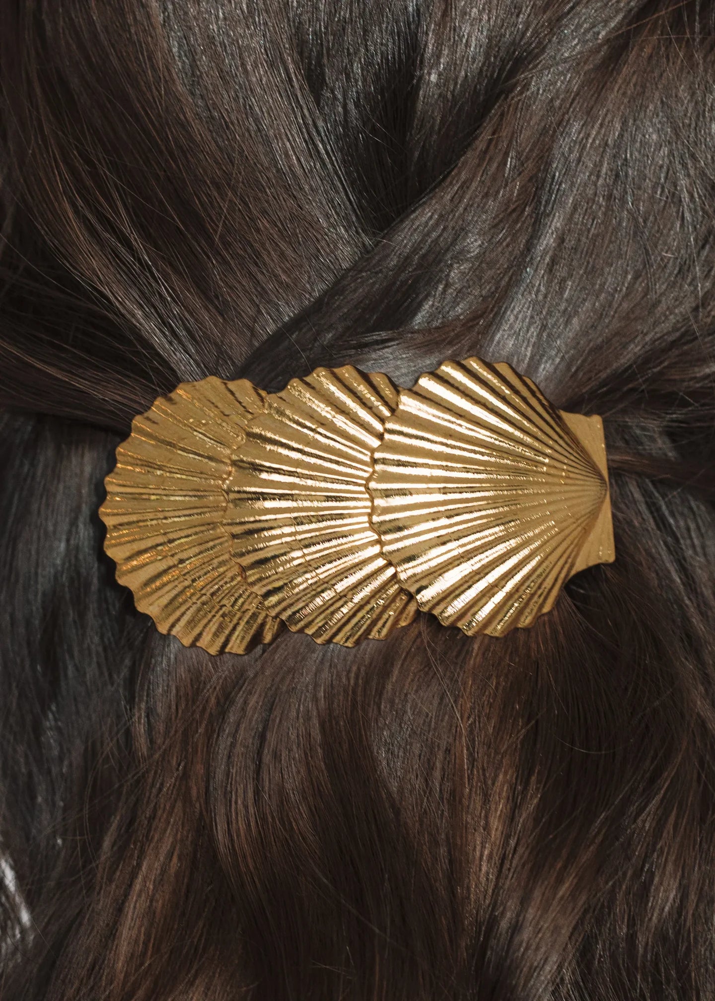 The Jennifer Behr Siren Barrette, shaped like two gold seashells, holds back dark brown hair securely with a French barrette clasp.
