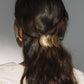 A person with long, wavy brown hair in a half-up style, secured with the Jennifer Behr Siren Barrette—an 18K gold accessory inspired by scallop shells—paired with a white top and gold earrings.