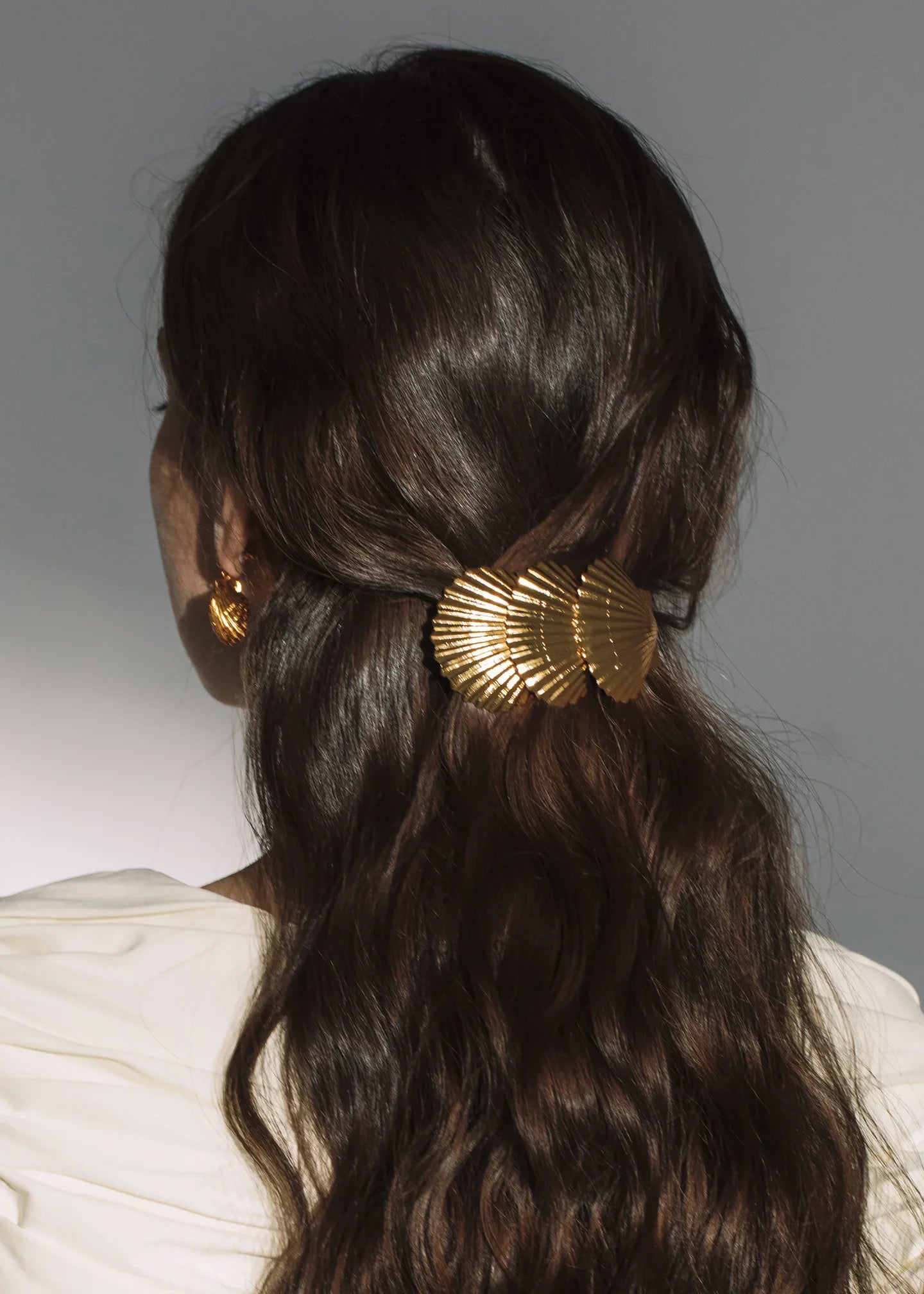 A person with long, wavy brown hair in a half-up style, secured with the Jennifer Behr Siren Barrette—an 18K gold accessory inspired by scallop shells—paired with a white top and gold earrings.