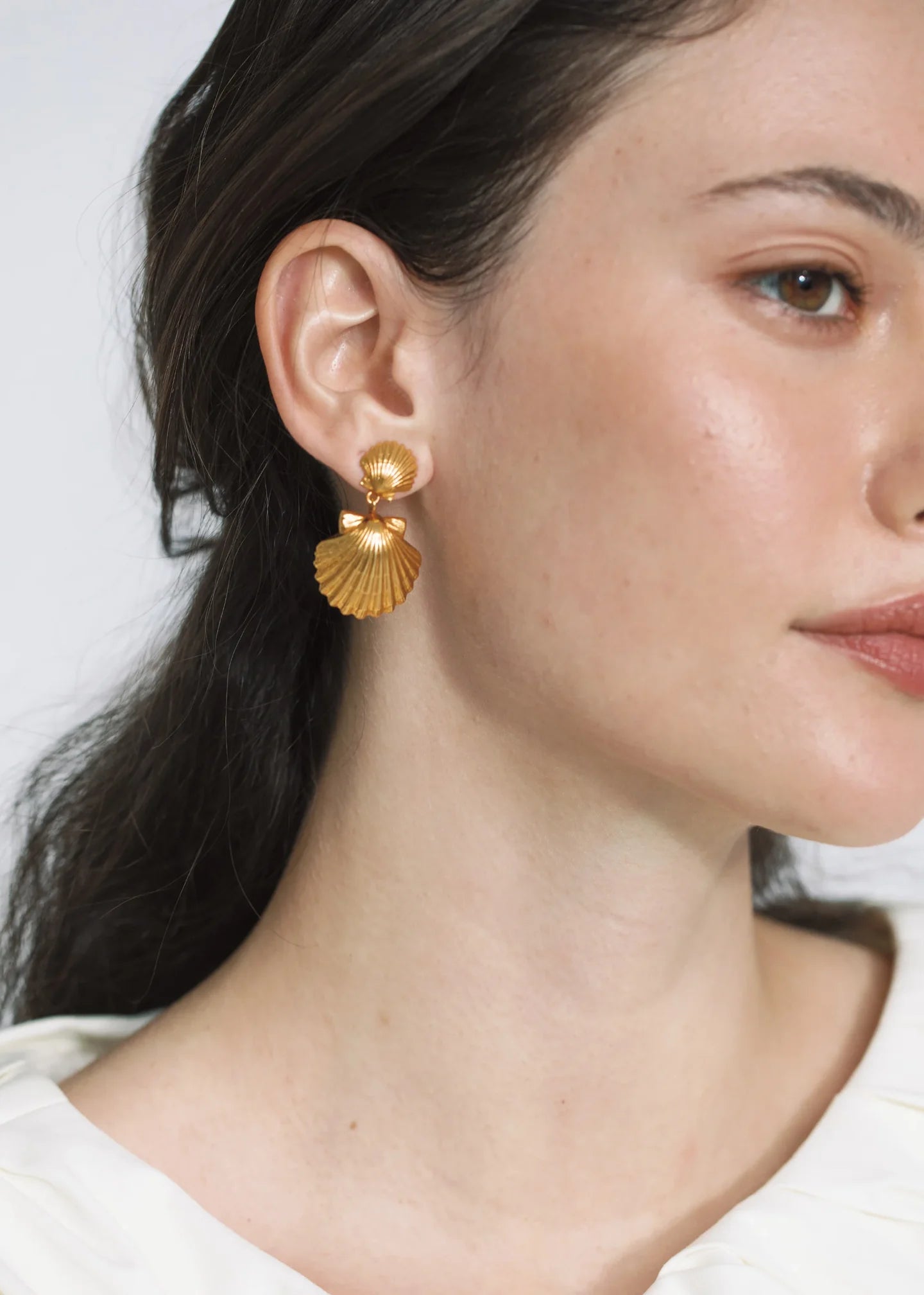 Close-up of a woman wearing Jennifer Behr's Caspian Earrings, crafted in 18-karat gold-plated metal with a seashell silhouette, her hair tucked behind her ear and a neutral expression.
