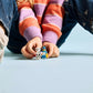 A child sits on the floor in striped socks, blue jeans, and a pink-striped sweater, holding the LEGO® Playground Fun with Bluey and Chloe set by Legos - Toyhouse along with another small figure.