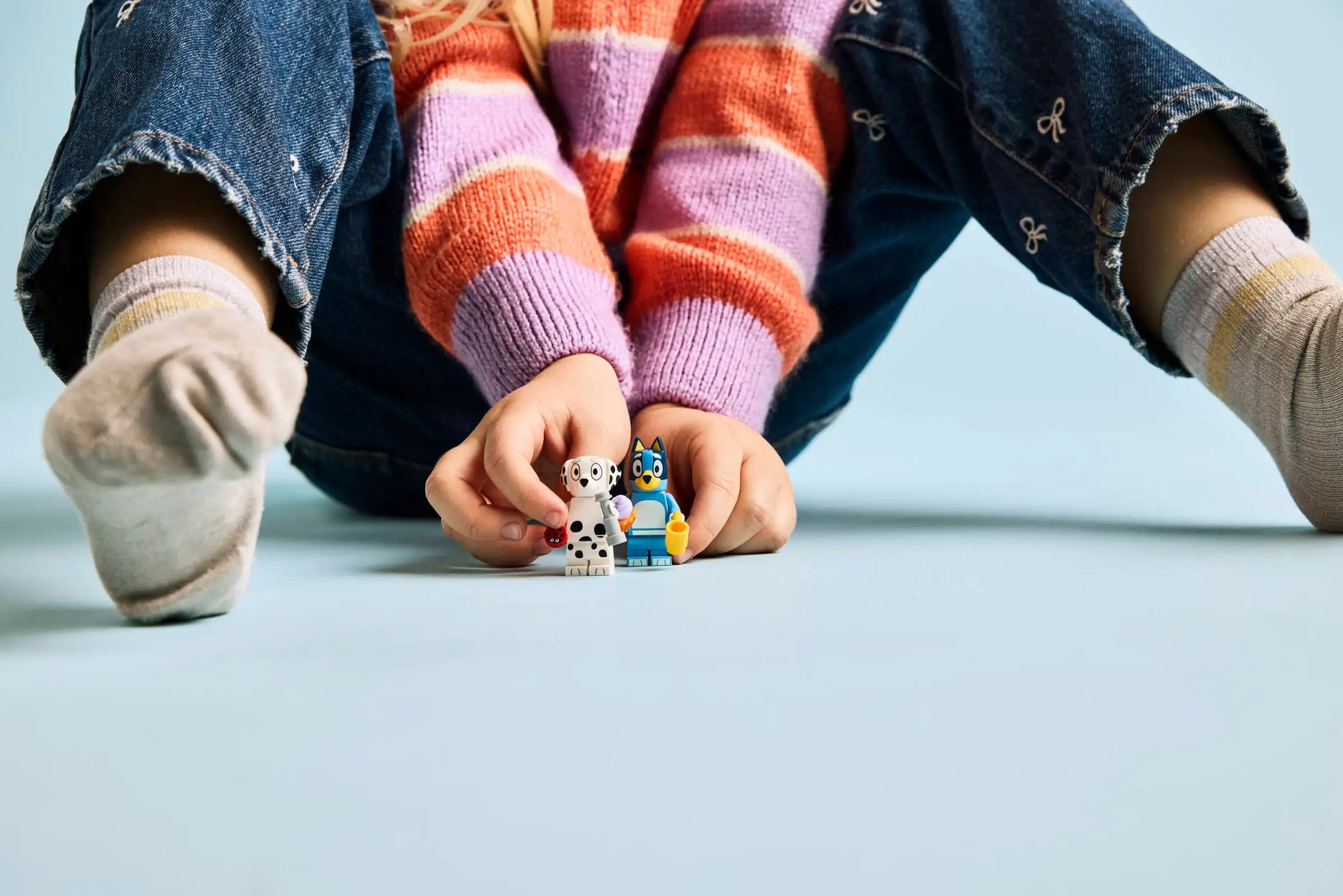 A child sits on the floor in striped socks, blue jeans, and a pink-striped sweater, holding the LEGO® Playground Fun with Bluey and Chloe set by Legos - Toyhouse along with another small figure.