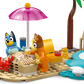 Two toy dog figures build a sandcastle with a pink umbrella, palm tree, red crab, and beach accessories in the LEGO® Bluey's Beach & Family Car Trip by Legos - Toyhouse—a playful preschool toy for imaginative adventures.