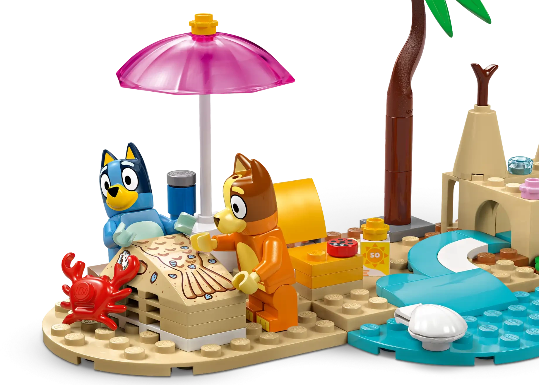 Two toy dog figures build a sandcastle with a pink umbrella, palm tree, red crab, and beach accessories in the LEGO® Bluey's Beach & Family Car Trip by Legos - Toyhouse—a playful preschool toy for imaginative adventures.