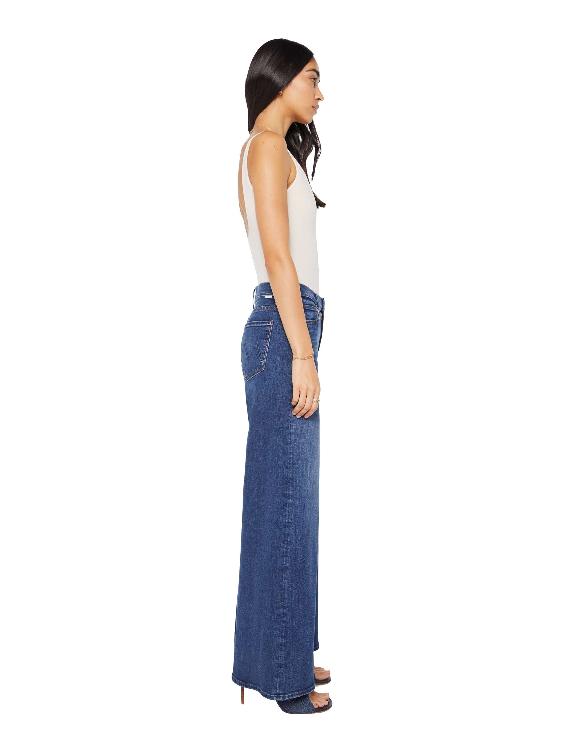 A woman stands in profile against a white background, wearing Mother’s The Undercover high rise wide leg jeans in recycled stretch denim, paired with a white sleeveless top and heeled sandals.