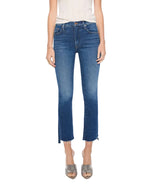 A person wearing a white top, Mother The Insider Ankle Step Fray high-waisted bootcut cropped jeans in stretch denim with a raw hem, and gray open-toe slide sandals, standing against a white background.