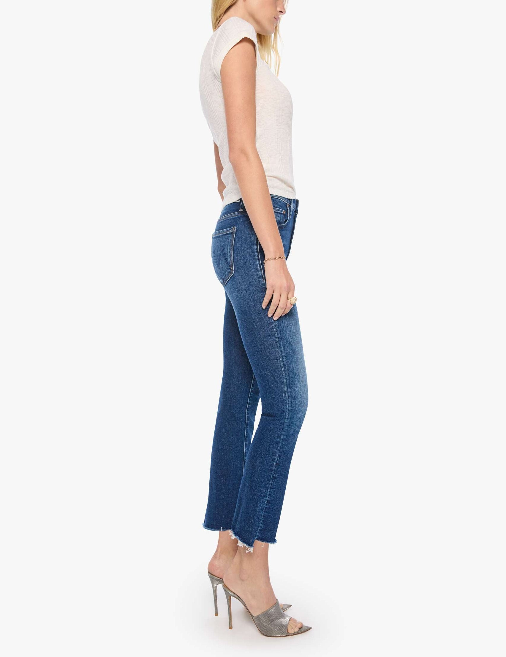 A woman stands in profile against a white background, wearing a short-sleeve white shirt, gray high-heeled shoes, and the Mother The Insider Ankle Step Fray high-waisted bootcut blue cropped jeans with frayed hems made from stretch denim.