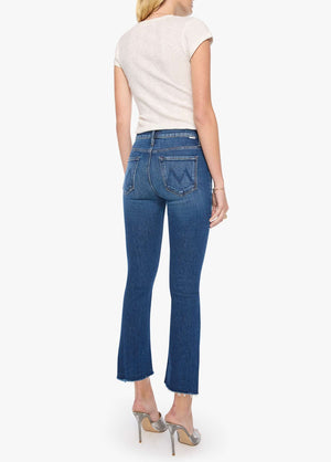 A woman stands with her back to the camera, wearing a short-sleeve beige shirt and Mother’s The Insider Ankle Step Fray blue cropped ankle jeans crafted from stretch denim, paired with silver high-heeled shoes.