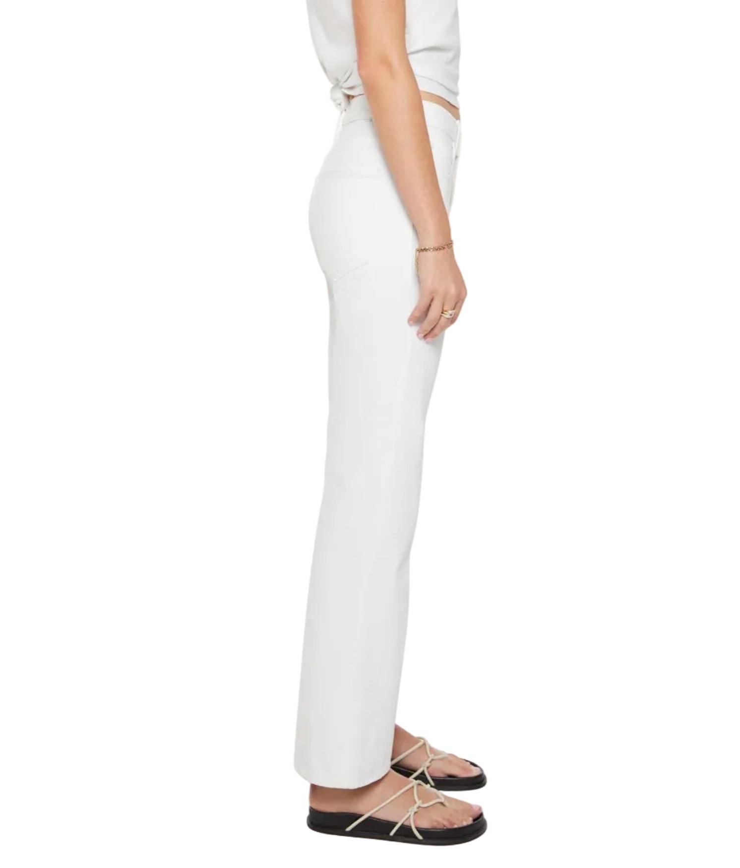 A woman stands in profile against a plain white background, wearing The Bookie Skimp high-waisted white denim pants by Mother, paired with a white crop top, black sandals, and a bracelet.