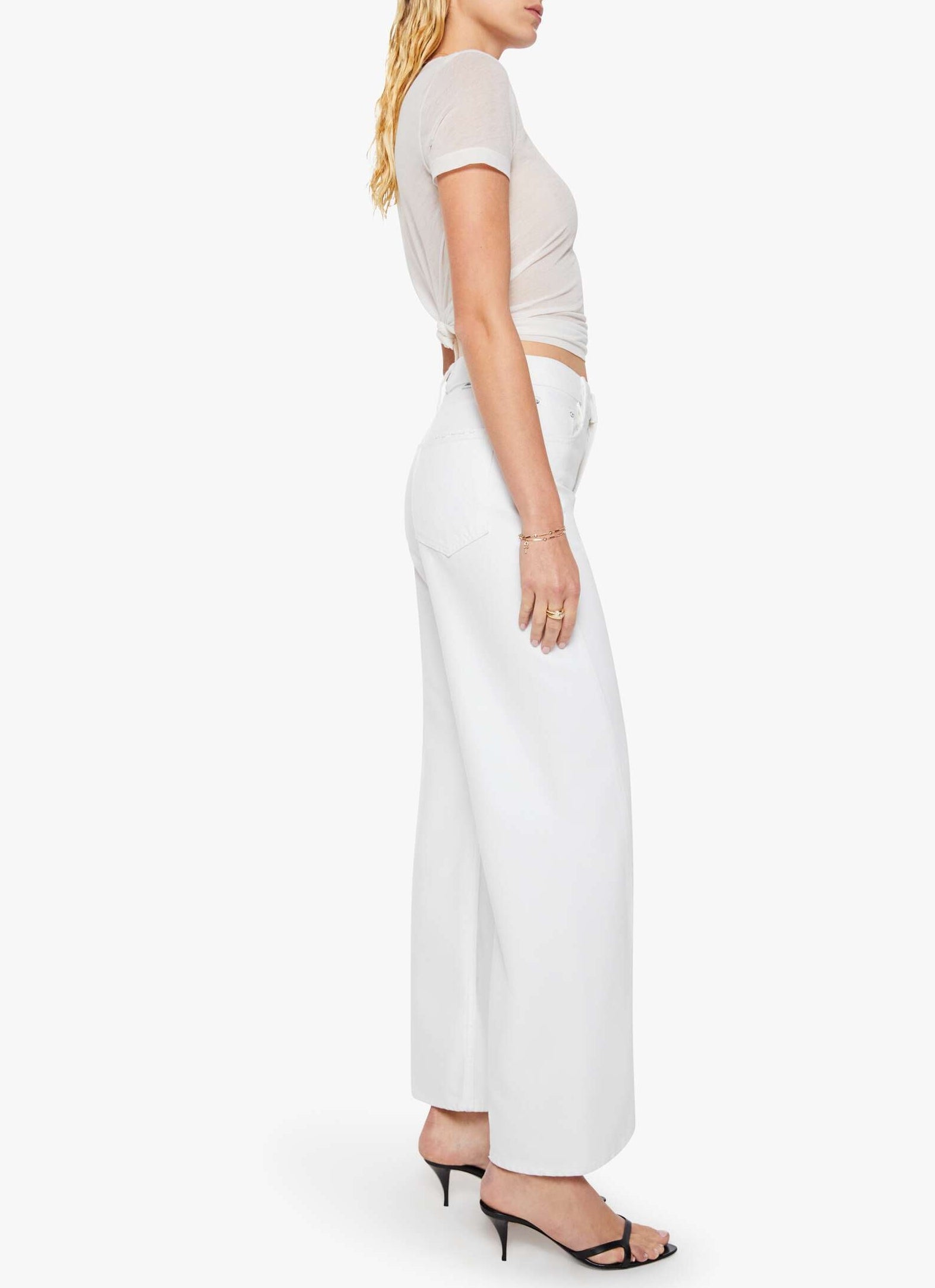 A woman stands in profile against a plain white background, wearing The Lemon Twist Skimp high-rise wide leg pants by Mother, styled with a sheer white t-shirt, black heeled sandals, and bracelets.