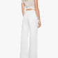 A woman is shown from the back wearing Mother’s The Lemon Twist Skimp wide-leg, high-rise white pants in a rigid cotton blend, paired with a sheer short-sleeve top. She has blonde wavy hair and black heeled sandals.