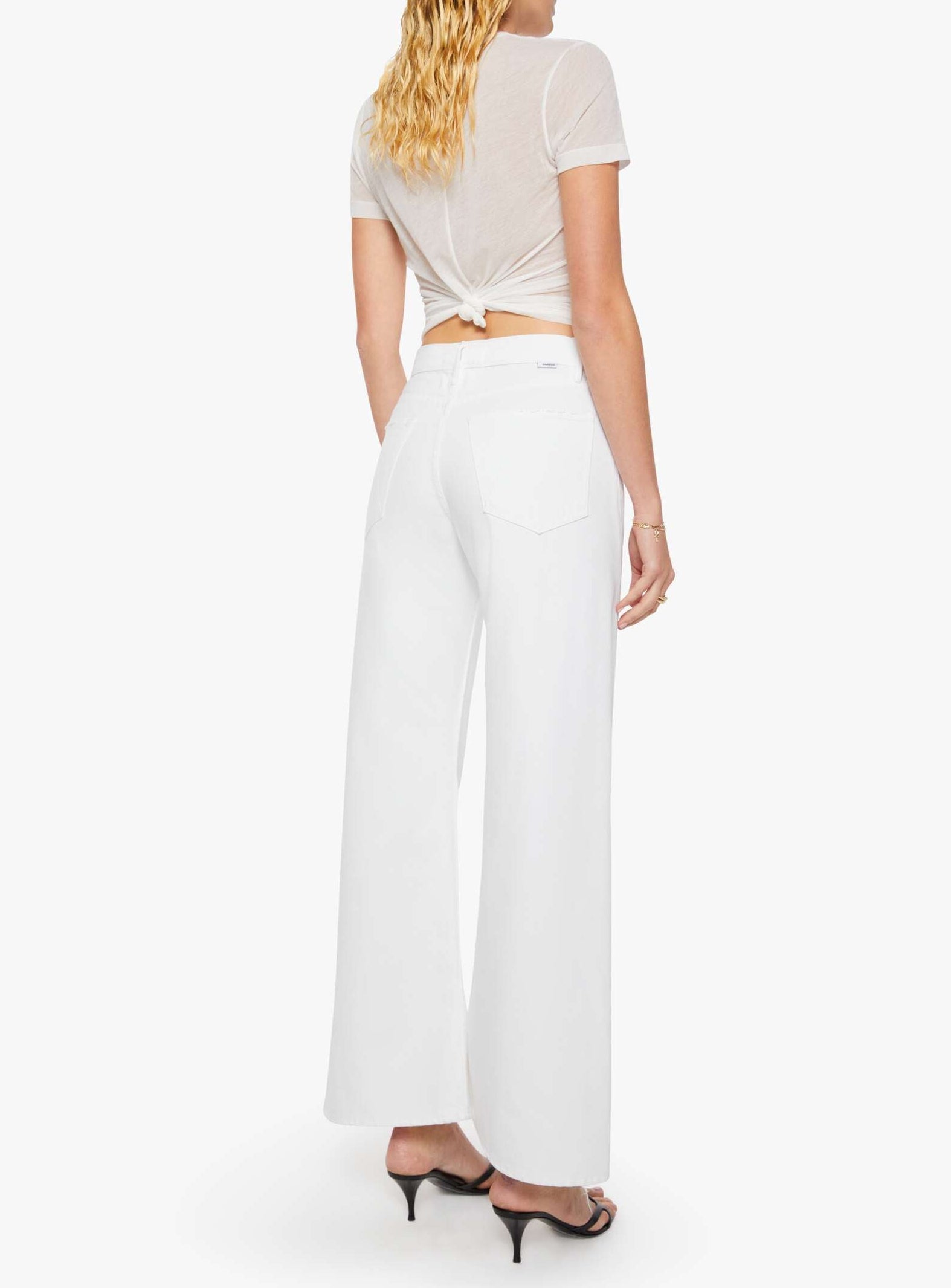 A woman is shown from the back wearing Mother’s The Lemon Twist Skimp wide-leg, high-rise white pants in a rigid cotton blend, paired with a sheer short-sleeve top. She has blonde wavy hair and black heeled sandals.