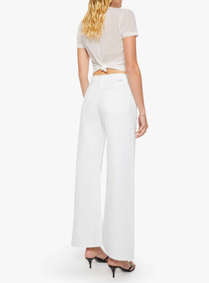 A woman is shown from the back wearing Mother’s The Lemon Twist Skimp wide-leg, high-rise white pants in a rigid cotton blend, paired with a sheer short-sleeve top. She has blonde wavy hair and black heeled sandals.