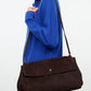 Wearing a bright blue sweater, this person carries the Staud Cy Oversized Bag—a dark brown suede shoulder bag featuring a flap closure and a long drop strap—creating a true statement piece.