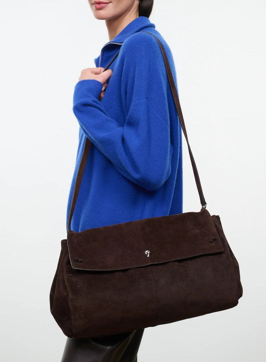 Wearing a bright blue sweater, this person carries the Staud Cy Oversized Bag—a dark brown suede shoulder bag featuring a flap closure and a long drop strap—creating a true statement piece.