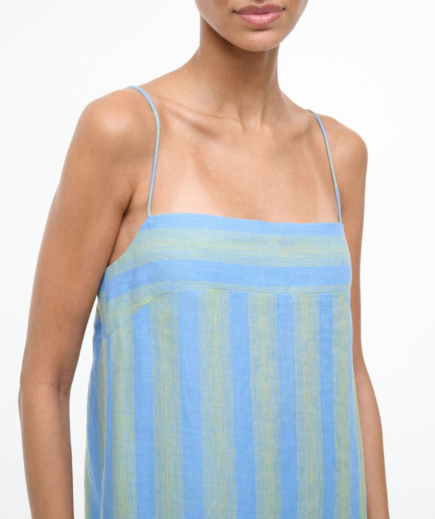 A person wearing the Staud Laura Linen Dress, featuring blue and green stripes and adjustable spaghetti straps, is shown from shoulders to mid-torso against a plain background.