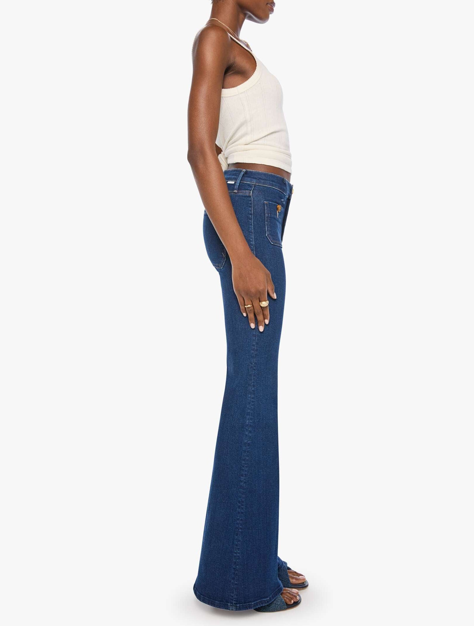 A woman stands in profile against a plain white background, wearing Mother’s The Patch Pocket Doozy high-rise flare jeans in dark blue stretch denim, paired with a sleeveless white top and sandals.