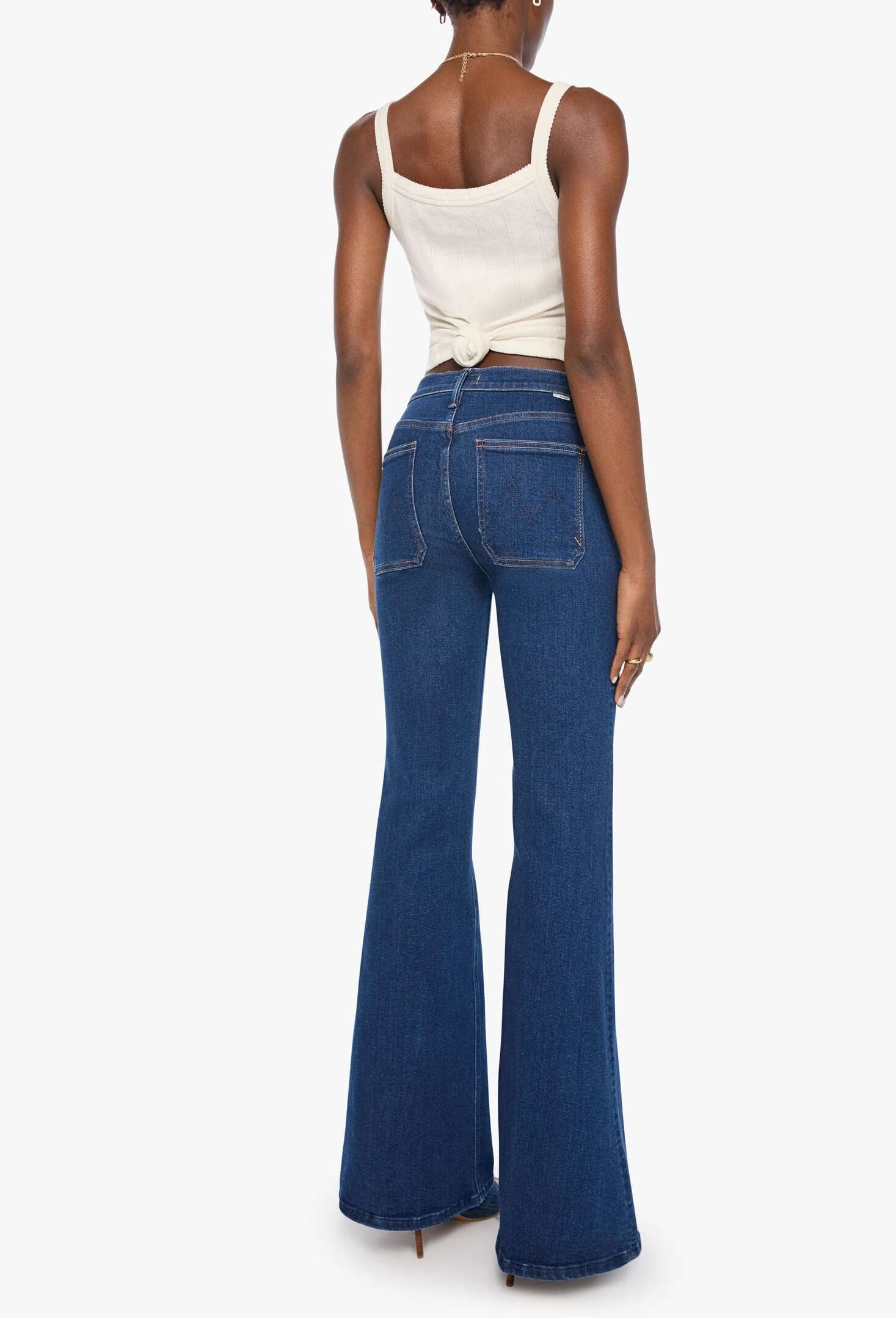 A person stands with their back to the camera, wearing Mother’s The Patch Pocket Doozy high-rise flare jeans in dark blue denim and a white sleeveless top, set against a plain white background.