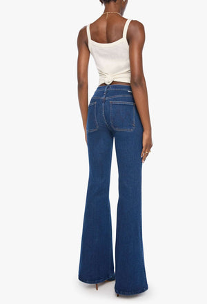 A person stands with their back to the camera, wearing Mother’s The Patch Pocket Doozy high-rise flare jeans in dark blue denim and a white sleeveless top, set against a plain white background.