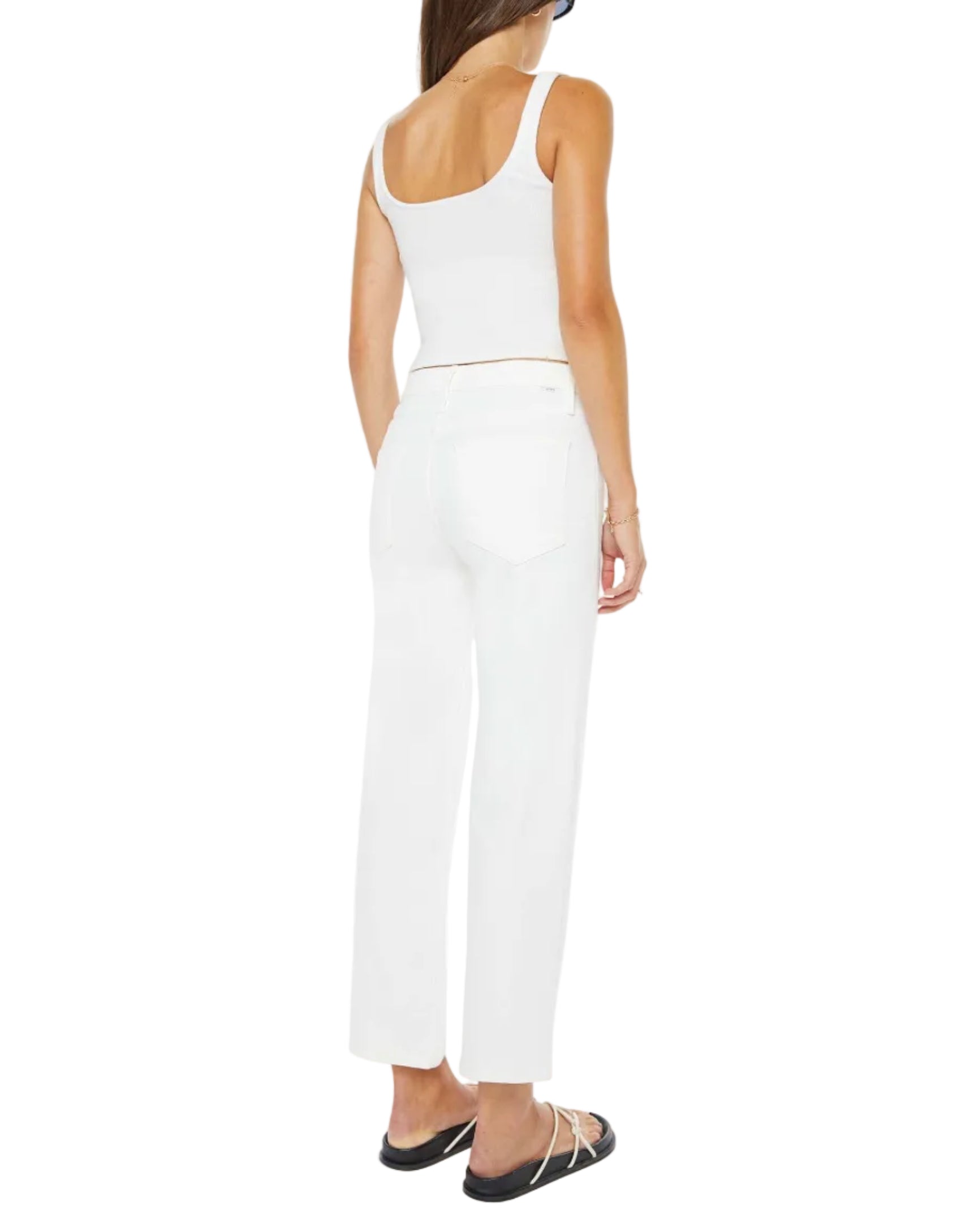 A woman faces away, wearing The Newbie Ankle high-rise stretch denim jeans from Mother, a white sleeveless crop top, black flat sandals, and a blue cap against a white background.