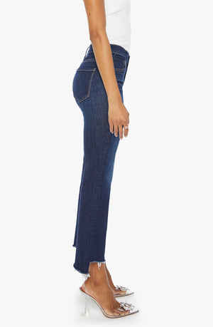 A person stands in profile wearing Mother’s The Insider Crop Step Fray high-waisted bootcut cropped stretch denim in dark blue, paired with a white sleeveless top and embellished clear heeled shoes.