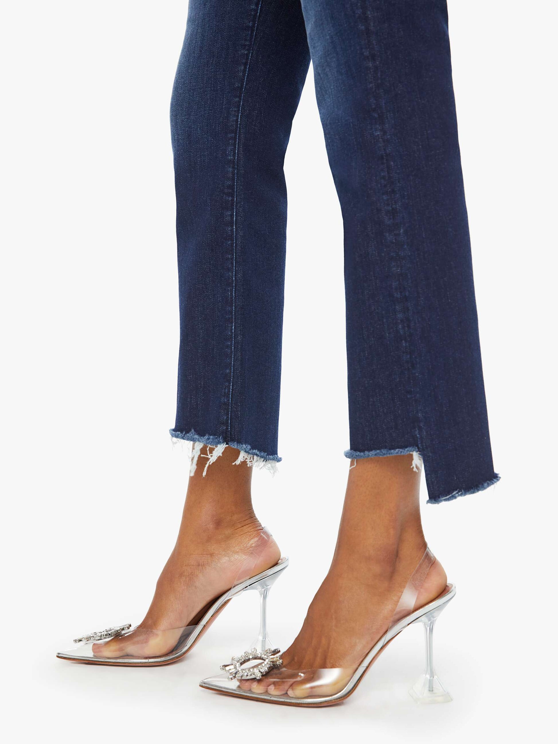 Someone wearing Mother’s The Insider Crop Step Fray high-waisted bootcut blue jeans with a frayed step-hem and clear, embellished pointed-toe heels, standing against a white background.
