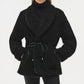 A person wears the Simkhai Kimia Tie Waist Jacket in black, featuring a textured fabric and tie front closure, over a black turtleneck and black pants with hands in the jacket pockets.