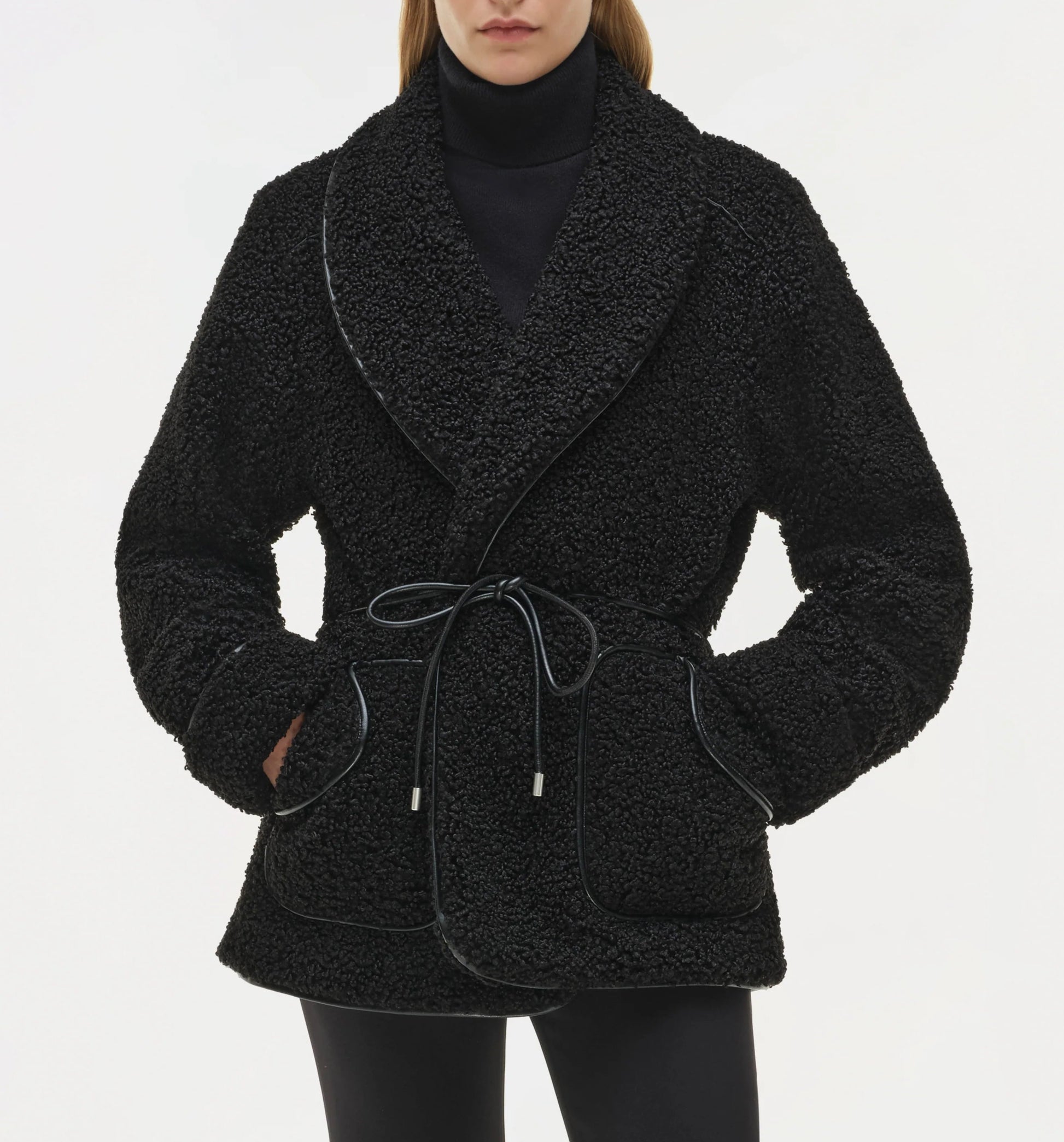 A person wears the Simkhai Kimia Tie Waist Jacket in black, featuring a textured fabric and tie front closure, over a black turtleneck and black pants with hands in the jacket pockets.