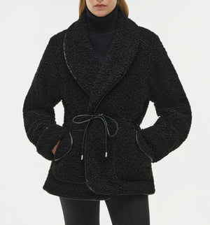 A person wears the Simkhai Kimia Tie Waist Jacket in black, featuring a textured fabric and tie front closure, over a black turtleneck and black pants with hands in the jacket pockets.
