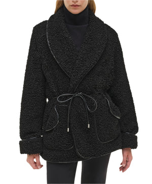 A person wearing the Simkhai Kimia Tie Waist Jacket—a black textured coat with a foldover collar, large lapels, front pockets, cuffed sleeves—paired with a black turtleneck and pants.