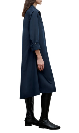 A person stands in profile against a white background wearing the TWP Hankerchief Midi Jenny's Gown, a navy blue long-sleeve cotton shirtdress, paired with black knee-high boots.