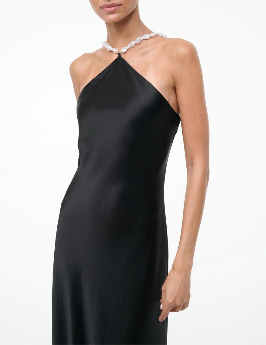 A woman wears the Staud Cadence Dress, a black satin slip with beaded pearl straps, shown against a plain white background.