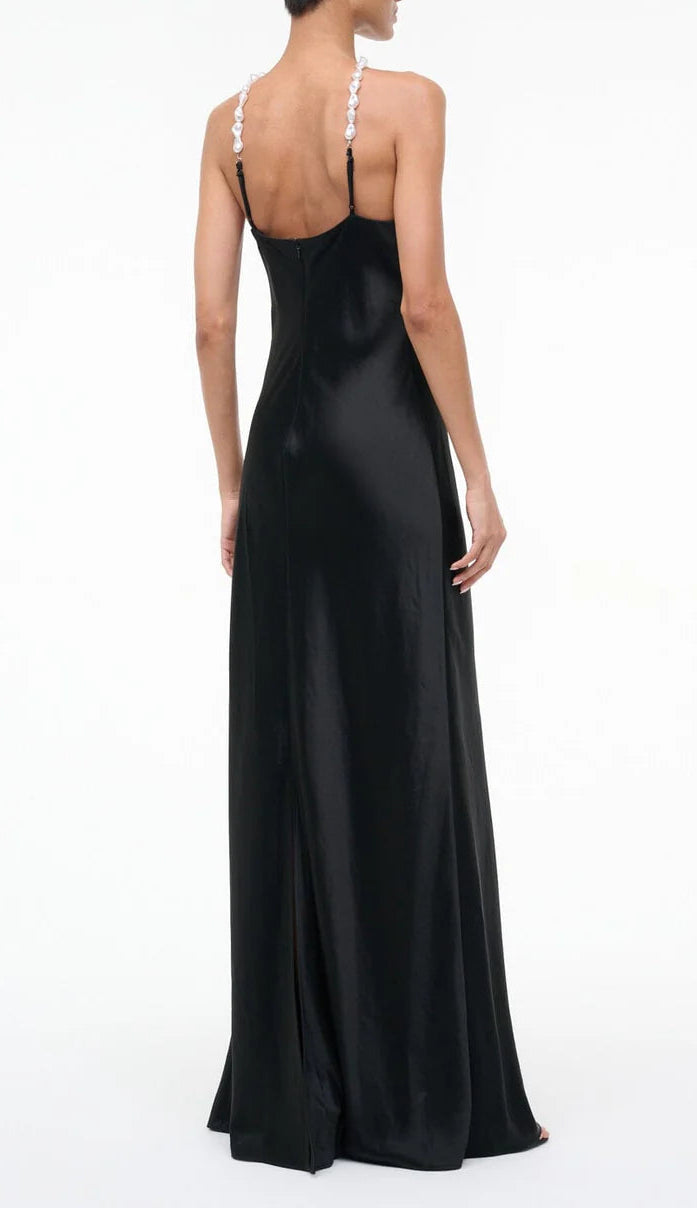 A woman stands facing away, wearing the Staud Cadence Dress—a long black satin slip with delicate beaded pearl straps and a subtle back slit.