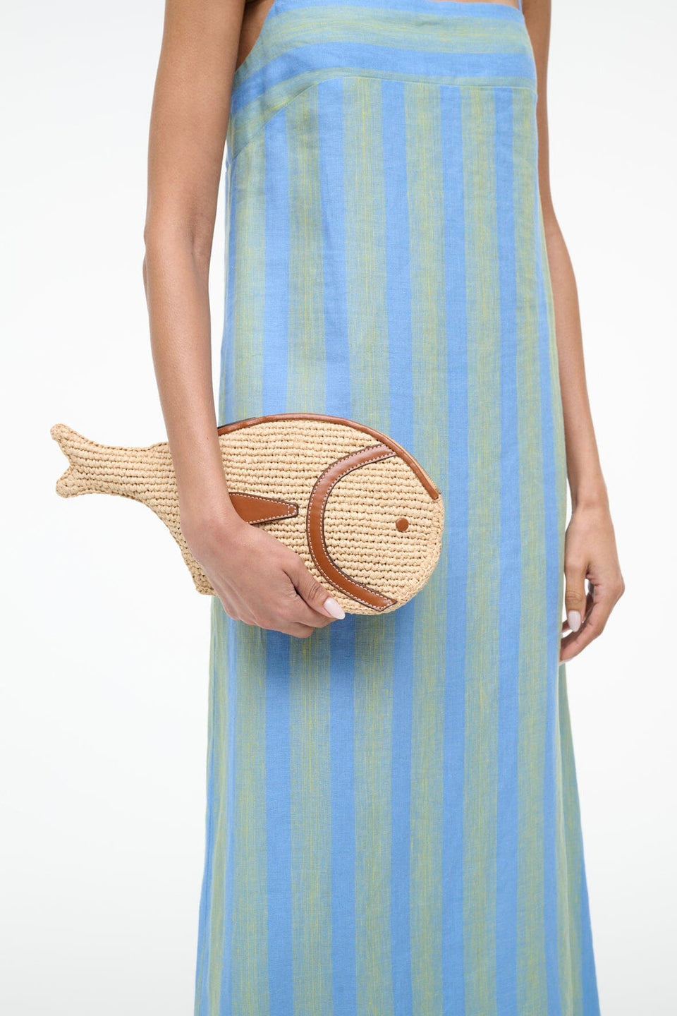 A person in a blue and green striped sleeveless dress holds the Staud Pesce Raffia Clutch, a fish-shaped bag crafted from natural raffia with leather trim.