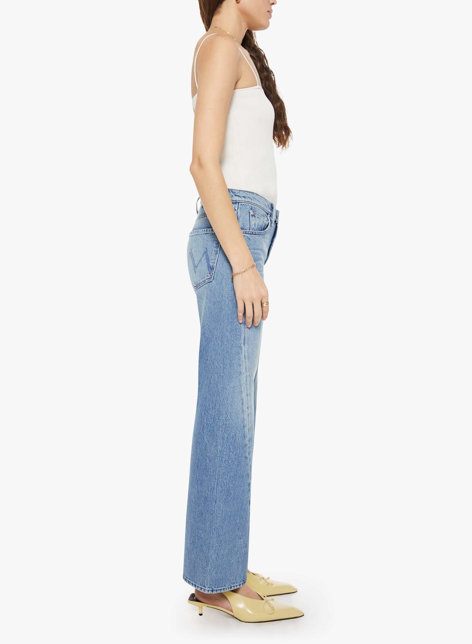 A woman stands in profile against a plain white background wearing a white tank top paired with Mother’s The Bookie Twist Skimp high-waisted light blue superior denim jeans and beige pointed-toe shoes.