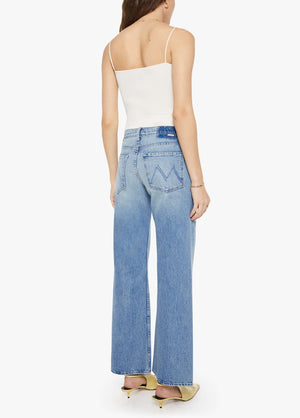 A woman stands with her back to the camera against a plain white background, wearing a white sleeveless top paired with Mother’s The Bookie Twist Skimp high-waisted, light blue bootcut jeans crafted from superior denim, and light-colored heels.