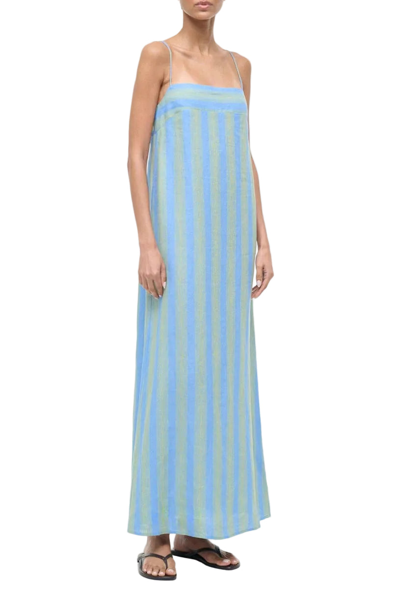 A person in the Staud Laura Linen Dress—a sleeveless, blue and green striped dress with adjustable spaghetti straps—paired with flip-flops, stands against a white background.