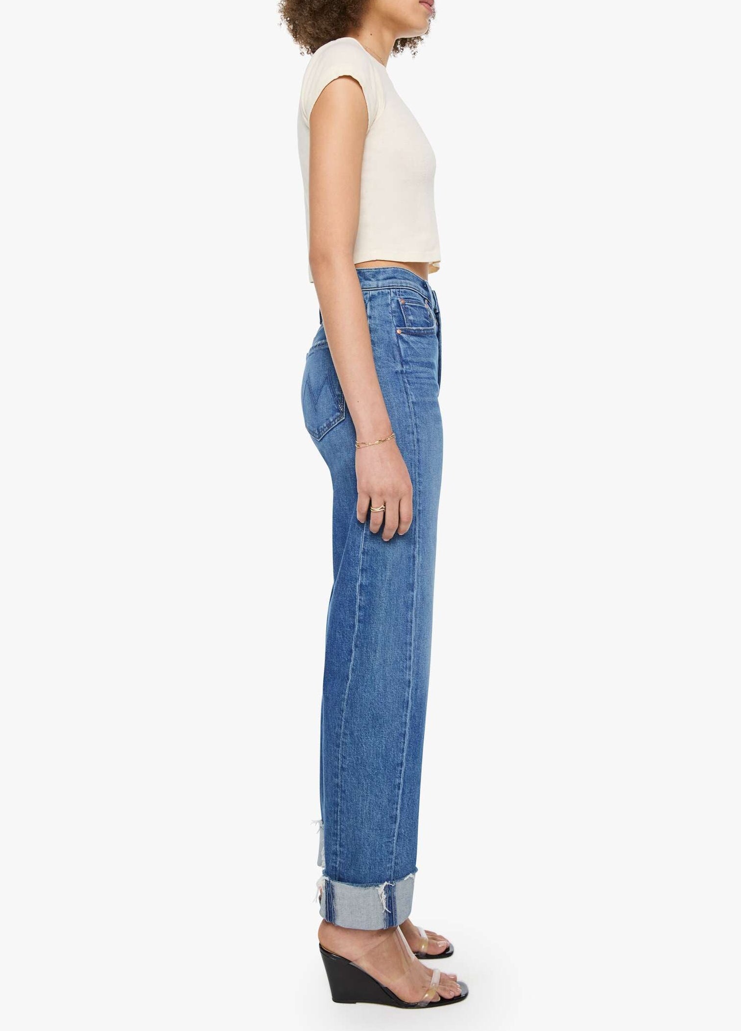 A woman stands in profile against a plain white background, wearing a short-sleeve white crop top, Mother’s Half Pipe Flood Cuff Fray high-waisted denim jeans with cuffed hems, and black wedge sandals.